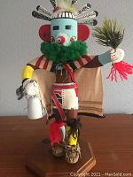 Full frontal view of the carved Hopi Kachina, showing detailed hand painting, feather headdress, green fluffy collar, and objects held in both hands, standing on an octagonal wooden base.