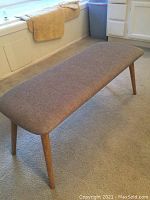 Photo showing angled view of rectangular gray upholstered bench with wooden tapered legs on carpeted floor.