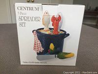 Box of Centrum 5-piece spreader set showing painted pot holder with vegetable decoration and spreaders with fish and lobster handles on lid
