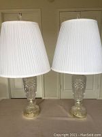 Pair of cut glass lamps with white pleated fabric shades, brass bases, and clear crystal bases with detailed cut glass design.