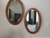 Two oval teak wood framed mirrors hanging on a wall, showing front reflective glass surfaces.