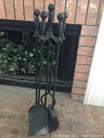 Full set of six wrought iron fireplace tools arranged in their matching stand, positioned in front of a brick fireplace.
