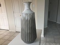 Photo of gray ceramic vase with vertical ribbed wave design, narrow neck and ombre finish on white surface against closed doors.