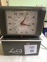 Front view of Lathem vintage time clock showing analog dial with 24-hour markings and red hands, textured gray metal case, and chrome logo.