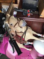 Light tan plastic toy horse with bridle and saddle, shown with assorted household items in background