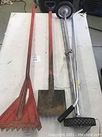 Three tools on a white table: two red-handled roofing shovels, one with a serrated blade and one with a flat square blade, alongside a chrome rolling magnet tool with rubber grips and wheels.