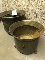 Two round decorative metal pots with hammered texture and lion head ring handles, one larger and one smaller. Visible rust inside and on surfaces.