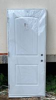 Front view of door wrapped in plastic showing two-panel design and knob/prep holes.