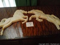 Two off-white ceramic horse wall decor pieces lying on a wooden surface, both showing side profiles in rearing positions.