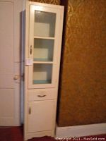 Full view of a tall white wood cabinet featuring one glass door on top with shelves, one drawer in the middle, and one bottom door for storage.
