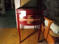 Photo of vintage side table with one drawer showing its red finish, brass hardware, and curved drawer front in an indoor setting.