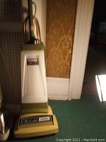 Photo of a cream and green Shetland heavy duty upright carpet shampooer resting upright near a wall with power cord and brush head visible.