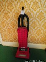 Red upright Dirt Devil vacuum cleaner with black hose leaning against a wall with yellow patterned wallpaper.