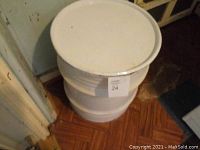 White steel drum container with removable top, standing on wooden floor near wall.