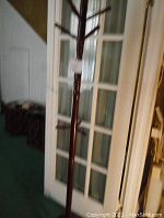 Full view of the tall wooden coat rack in a room with green carpet and glass-paneled door in the background. Shows the condition and visible missing hook.