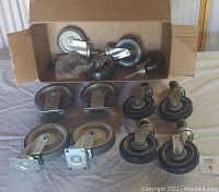 Photo shows a variety of 11 caster wheels in a mix of brands and styles, arranged on a cloth surface with some in a cardboard box in the background. Wheels measure 5 to 6 inches diameter with metal brackets.