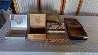 Photo showing six assorted cigar boxes including metal and wooden boxes of various sizes and styles displayed on table