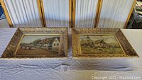 Two framed oil paintings side by side on table showing rural cottage scenes