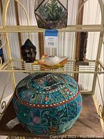 Shelf displaying round embroidered ottoman, wooden bust figurines, and glass bird figurine on decorative plate.