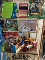 Photo showing Leapfrog LeapPad, Speak & Spell, Camp Aura Outdoor Adventure kit on top shelf; Bop It game, Star Wars drip toy and learning block toys on low shelves.