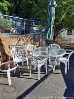 Full view of patio table with umbrella and six white plastic chairs plus extra white plastic chair and side table outside in daytime.