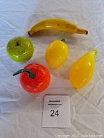 Top view of all five hand blown glass fruits on a white cloth background showing banana, apple, lemon, orange, and pear shapes.