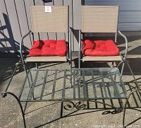 Two beige frame patio chairs with red cushions and black metal frame coffee table with glass top shown outside on concrete.