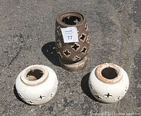 Photo of three tabletop candle clay pots: two smaller white round pots and one larger brown cylindrical pot with decorative cutouts, shown outdoors on a paved surface.