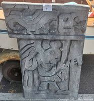 Grey faux concrete panel with carved Aztec scene leaning against a white surface outdoors.