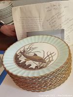 Stack of the 12 porcelain plates showing scalloped gilt edges from Edwin J.D. Bodley with some paperwork in background.