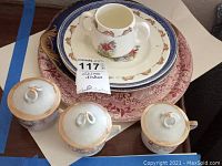 Photo showing child's Royal Doulton Bunnykins plate and cup set stacked on Blue Willow and Spanish Festivities plates with three lusterware cups in front