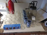 Various glassware and kitchen items arranged on countertop including glass butter churn, blue grape design Hazel Atlas water tumblers, and mid-century clear glass goblets.