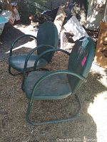 Pair of green metal lawn chairs sitting outside on grass and dirt, showing surface rust and wear.
