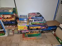 Stacked board games showing titles Orient Express, Checkers, Word Power, Crossfire, Battle Cry, Pathfinder, Dune, and others on shelves and floor, boxes visible with wear