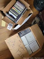 Two boxes on a wooden floor, one filled with assorted CDs and the other with empty cases.