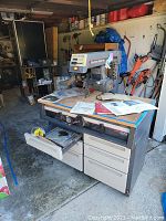 Wide view of Sears Craftsman radial arm saw on metal stand with drawers, mobile on wheels with a brake, and several manuals and papers on the wooden table surface.