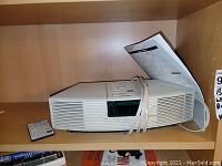 Bose Wave Radio unit on wooden shelf with power cord and instruction manual resting on top, remote control placed beside it.