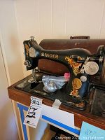 Black vintage Singer hand crank sewing machine on wooden table with original wood dome case behind it. Visible bobbin, shuttle, needles in a plastic bag on machine base. Machine shows gold and red ornate decals and some wear.