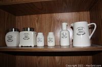 Photo showing entire set of six vintage pottery condiment containers arranged on a wooden shelf. Includes labeled jars for sugar, tea, vinegar, salt, pepper, and a milk jug.