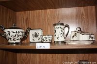 Photo of four ceramic items on wooden shelf showing teapot, sugar bowl, creamer with lid, and butter dish. Each item has silver or platinum lustre bands and leaf decoration on cream base.