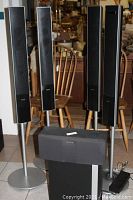Photo showing four tall speakers on stands plus center speaker and subwoofer arranged in front of wooden chairs and other furniture.