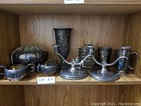 Photo of a collection of silver plated items including candelabras, mugs, cream and sugar holders, a decorative frog figurine, and a tall vase on a wooden shelf.