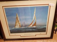 Framed print showing two sailing yachts with sails on the water under a blue sky with a lighthouse in the background, with title plaque visible.