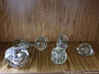 Photo of six clear blown glass paperweights on wooden surface, showcasing bubble inclusions and various organic shapes including pumpkin and apple.