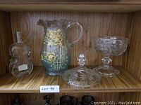 Wooden shelf displaying four vintage glass items: a Moser pitcher with gold floral decoration, a violin-shaped glass decanter, a covered cut crystal dish, and a footed crystal compote bowl.