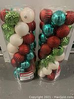 Two large clear plastic containers each containing 50 shatter-proof Christmas ornaments in red, green, teal, and white colors with various finishes.