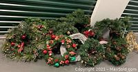 Photo of 13 assorted green Christmas wreaths laid out together, showing variations in size and decoration including red ornaments, berries, pinecones, and some white twig styled wreaths.