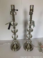 Pair of clear acrylic and metal lamp bases without bulbs, standing side by side against a plain white background, showing tags attached to each lamp.