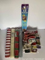 Photo showing Deluxe EZ Bow Maker in original vertical packaging and assortment of ribbon rolls including four large mesh rolls and multiple smaller ribbon spools with diverse colors and patterns.
