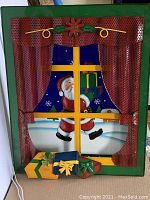 Front view of the metal light-up sign showing Santa Claus and gifts outside a window with light bulbs and red curtains.